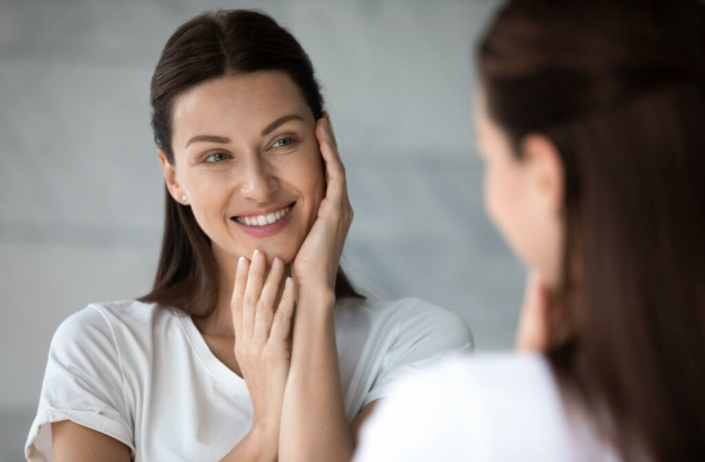 A person smiles as they touch their skin while looking in the mirror.