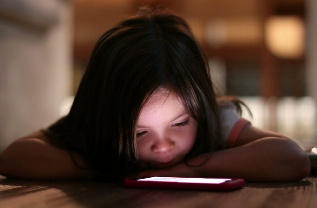 A child lies on the floor in a dim room, closely watching a glowing tablet screen.