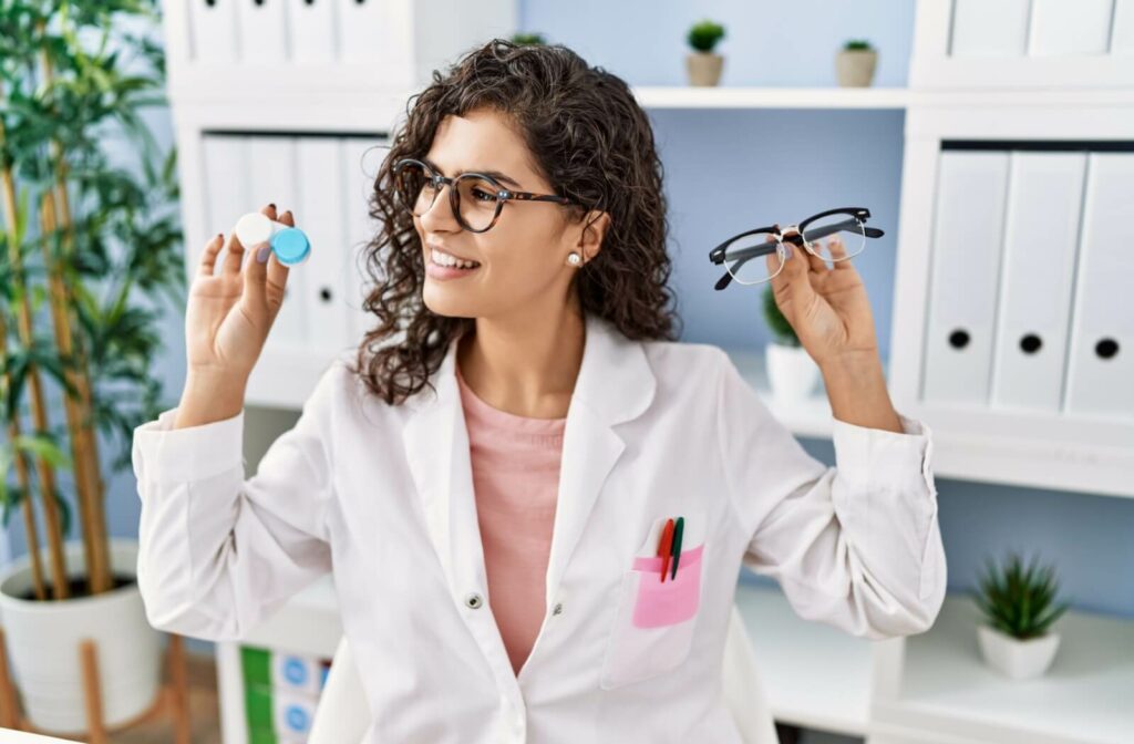 A young optometrist with curly hair holds up a pair of eyeglasses in one hand and a contact lens case in the other and turns their head to look at the contact lens case.